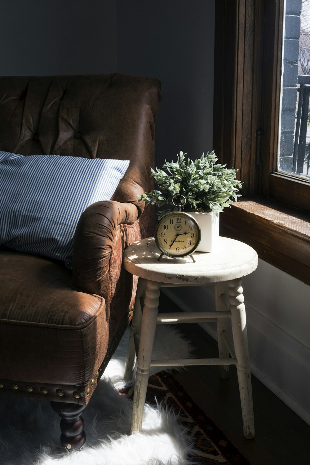 Poltrona da divano imbottita in pelle marrone accanto alla sedia sgabello in legno bianco con orologio analogico rotondo in metallo grigio