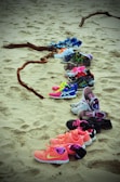 A collection of various colorful athletic shoes arranged in a line on sandy ground, with pieces of driftwood scattered nearby. The shoes display a variety of bright colors including pink, blue, neon green, and orange, suggesting a casual and informal setting.