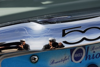 A reflection on a shiny metallic surface displaying two people holding cameras, with the number 50 partially visible. The surface also reflects a blue and white sign with the words 'Beautiful Ohio 1803.'