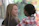 Two individuals are interacting, with one person appearing to be speaking while the other listens attentively. The background is softly blurred, displaying some colorful abstract patterns.