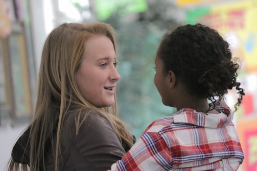 Two individuals are interacting, with one person appearing to be speaking while the other listens attentively. The background is softly blurred, displaying some colorful abstract patterns.