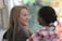 Two individuals are interacting, with one person appearing to be speaking while the other listens attentively. The background is softly blurred, displaying some colorful abstract patterns.