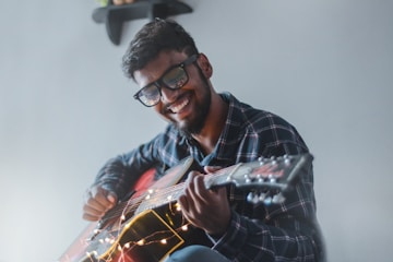 A person wearing glasses is playing an acoustic guitar with strings decorated by fairy lights. They are smiling joyfully and wearing a plaid shirt. The background is simple, with a small shelf visible in the upper corner.