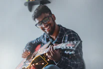 A person wearing glasses is playing an acoustic guitar with strings decorated by fairy lights. They are smiling joyfully and wearing a plaid shirt. The background is simple, with a small shelf visible in the upper corner.