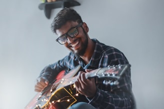A person wearing glasses is playing an acoustic guitar with strings decorated by fairy lights. They are smiling joyfully and wearing a plaid shirt. The background is simple, with a small shelf visible in the upper corner.
