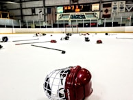 An indoor ice hockey rink with various pieces of hockey equipment scattered on the ice, including helmets and sticks. Banners and flags adorn the walls above the rink, and a scoreboard displays the end of a game.