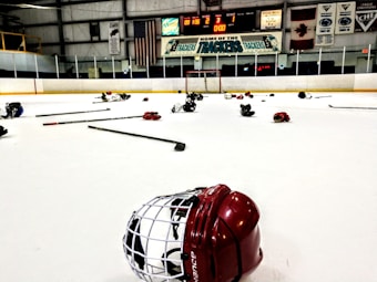 An indoor ice hockey rink with various pieces of hockey equipment scattered on the ice, including helmets and sticks. Banners and flags adorn the walls above the rink, and a scoreboard displays the end of a game.