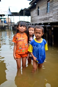Three young children stand in knee-deep water in front of wooden stilt houses. The child on the left wears an orange outfit, the one in the middle wears a blue and yellow T-shirt, and the child in the back has a partially visible shirt. The children are barefoot, and the water they stand in is murky. The background consists of wooden structures and stilts, suggesting a waterside or flood-prone location.