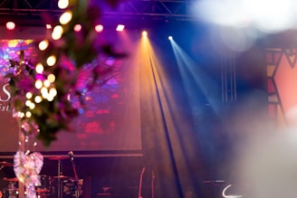 An indoor stage setup with colorful stage lights casting blue and yellow beams. The foreground features out-of-focus hanging lights and decoration that appears floral. The backdrop consists of abstract, geometric patterns in vibrant colors, and a drum set is visible on the stage.