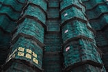 Tall structures covered in green construction netting with scaffolding. There are signs with text, possibly in an East Asian script, attached to the netting.