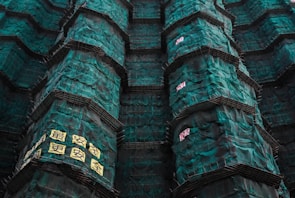 Tall structures covered in green construction netting with scaffolding. There are signs with text, possibly in an East Asian script, attached to the netting.