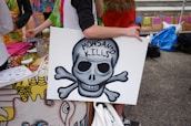 A person is holding a sign featuring a skull and crossbones with the words 'Monsanto Kills' written on it. The background includes a colorful tablecloth and various items such as a cup and papers, suggesting an outdoor event or protest setting. Several people are partially visible, engaging with the scene.