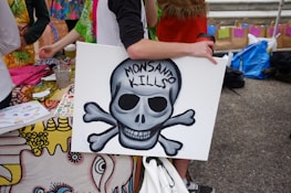 A person is holding a sign featuring a skull and crossbones with the words 'Monsanto Kills' written on it. The background includes a colorful tablecloth and various items such as a cup and papers, suggesting an outdoor event or protest setting. Several people are partially visible, engaging with the scene.