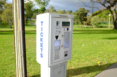 A ticket vending machine is positioned in a park surrounded by green grass and trees under a cloudy sky. The machine has visible buttons, a display screen, and signage with the word 'TICKETS' printed vertically on its side.