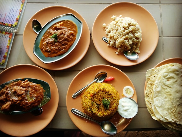 A variety of Indian dishes are arranged on round, orange plates. The dishes include two servings of a spicy curry in oval metal dishes, a mound of rice, and biryani accompanied by a small serving of yogurt and a slice of lime. A stack of flatbreads is to the right. Spoons are placed on the plates, and a menu is partially visible to the side.