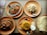 A variety of Indian dishes are arranged on round, orange plates. The dishes include two servings of a spicy curry in oval metal dishes, a mound of rice, and biryani accompanied by a small serving of yogurt and a slice of lime. A stack of flatbreads is to the right. Spoons are placed on the plates, and a menu is partially visible to the side.