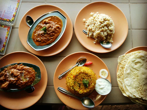 A variety of Indian dishes are arranged on round, orange plates. The dishes include two servings of a spicy curry in oval metal dishes, a mound of rice, and biryani accompanied by a small serving of yogurt and a slice of lime. A stack of flatbreads is to the right. Spoons are placed on the plates, and a menu is partially visible to the side.