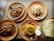 A variety of Indian dishes are arranged on round, orange plates. The dishes include two servings of a spicy curry in oval metal dishes, a mound of rice, and biryani accompanied by a small serving of yogurt and a slice of lime. A stack of flatbreads is to the right. Spoons are placed on the plates, and a menu is partially visible to the side.