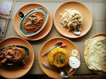 A variety of Indian dishes are arranged on round, orange plates. The dishes include two servings of a spicy curry in oval metal dishes, a mound of rice, and biryani accompanied by a small serving of yogurt and a slice of lime. A stack of flatbreads is to the right. Spoons are placed on the plates, and a menu is partially visible to the side.