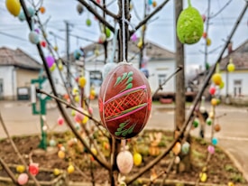 A decorative Easter egg hangs from a tree branch, intricately painted with colorful patterns. In the background, more similar eggs are attached to bare branches, suggesting a festive or springtime celebration. Houses and a street can be seen out of focus.