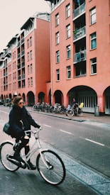 A woman wearing sunglasses, a dark jacket, and ripped jeans is cycling on a white bicycle along a street lined with a row of red apartment buildings. Several other bicycles are parked along the buildings, and there is a person in a yellow jacket in the background near the entrance of one of the buildings.