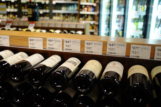 A wine store shelf containing various bottles of wine with price tags above them. The background shows refrigerator units with illuminated glass doors displaying more bottles. Labels on the wine bottles are partially visible, with different price points displayed on the shelf.