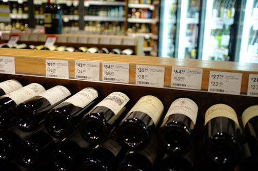 A wine store shelf containing various bottles of wine with price tags above them. The background shows refrigerator units with illuminated glass doors displaying more bottles. Labels on the wine bottles are partially visible, with different price points displayed on the shelf.