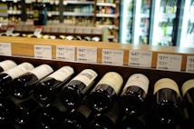 A wine store shelf containing various bottles of wine with price tags above them. The background shows refrigerator units with illuminated glass doors displaying more bottles. Labels on the wine bottles are partially visible, with different price points displayed on the shelf.