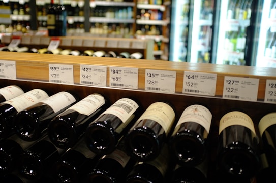 A wine store shelf containing various bottles of wine with price tags above them. The background shows refrigerator units with illuminated glass doors displaying more bottles. Labels on the wine bottles are partially visible, with different price points displayed on the shelf.