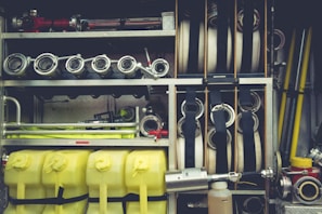 A neatly organized collection of firefighting equipment, including coiled hoses, metal nozzles, yellow plastic containers, and various metal pipes and fittings. The equipment is stored within compartments on a fire truck, showcasing its readiness and accessibility.