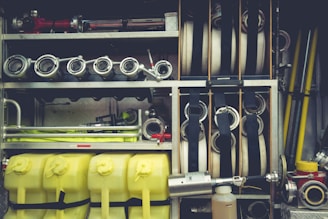 A neatly organized collection of firefighting equipment, including coiled hoses, metal nozzles, yellow plastic containers, and various metal pipes and fittings. The equipment is stored within compartments on a fire truck, showcasing its readiness and accessibility.
