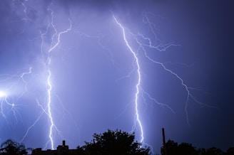 Dramatic lightning bolts illuminate a dark sky, casting bright, jagged lines across the scene. Silhouetted trees and rooftops are visible at the bottom of the image, adding depth and context to the threatening weather scenario.
