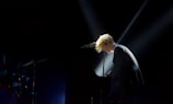 A musician with blonde hair is intensely playing a grand piano on stage. The scene is dimly lit, with a spotlight focused on the performer, creating a dramatic and intimate atmosphere. The background is mostly dark, highlighting the musician and the piano. There is a drum set partially visible to the left.
