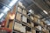 A warehouse scene with tall metal shelves stacked with cardboard boxes and pallets. The structure has a high ceiling with visible panels and lighting fixtures. Signs labeled 'Rack 2' and 'Rack 4' are prominently displayed.