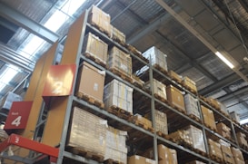 A warehouse scene with tall metal shelves stacked with cardboard boxes and pallets. The structure has a high ceiling with visible panels and lighting fixtures. Signs labeled 'Rack 2' and 'Rack 4' are prominently displayed.