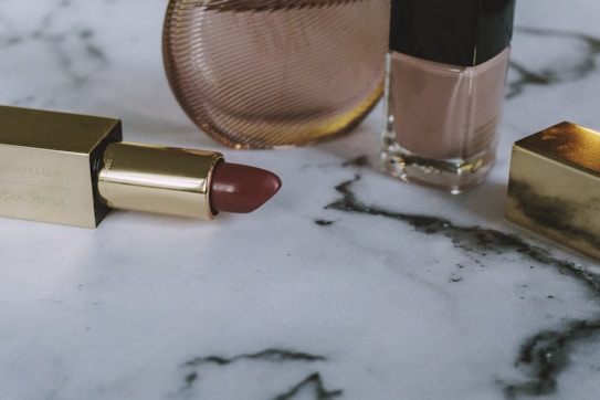 A close-up of a gold-cased lipstick lying on a marble surface alongside a bottle of nude nail polish and a round compact with a textured surface, all arranged aesthetically.
