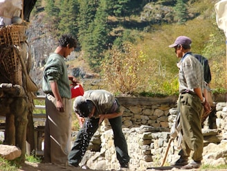 Three men are outdoors in a rural, mountainous setting. One man is pouring water from a red container onto another man's head, presumably for washing or cooling off. The third man is observing. They are surrounded by stone structures and greenery, suggesting an agricultural or remote area.