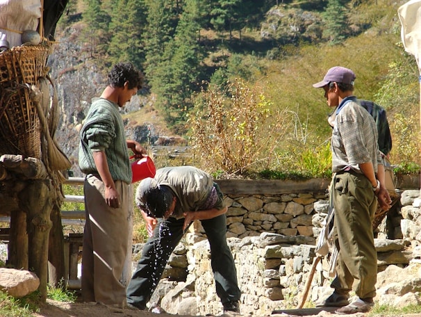 Three men are outdoors in a rural, mountainous setting. One man is pouring water from a red container onto another man's head, presumably for washing or cooling off. The third man is observing. They are surrounded by stone structures and greenery, suggesting an agricultural or remote area.