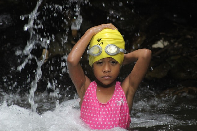 A young child stands in water, wearing a yellow swim cap and goggles on their forehead. The child is dressed in a pink swimsuit with white patterns and is looking calmly at the camera. Water splashes around them as they hold their arms behind their head.