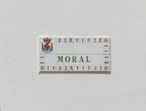 A ceramic tile sign with the word 'MORAL' prominently displayed in green letters. The sign is bordered with small symbols in brown and features an ornate crest with a crown and foliage on the top left corner.
