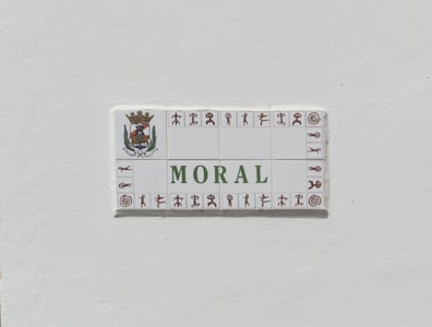 A ceramic tile sign with the word 'MORAL' prominently displayed in green letters. The sign is bordered with small symbols in brown and features an ornate crest with a crown and foliage on the top left corner.