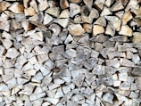 A tightly stacked pile of chopped firewood, with the cut ends of the logs facing outward. The wood varies in shades of brown and gray, with visible textures and grain patterns.