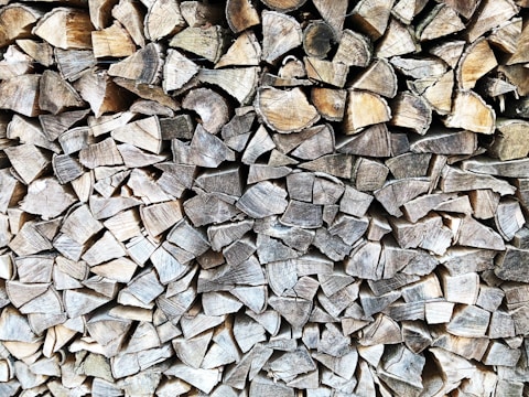 A tightly stacked pile of chopped firewood, with the cut ends of the logs facing outward. The wood varies in shades of brown and gray, with visible textures and grain patterns.