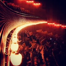 An auditorium with wooden interior features dimly lit, warm lights casting an amber glow. The seating area has several people scattered throughout, some are seated while others are standing or conversing. The curved lights run parallel to the rows of seats creating an elegant and intimate atmosphere.