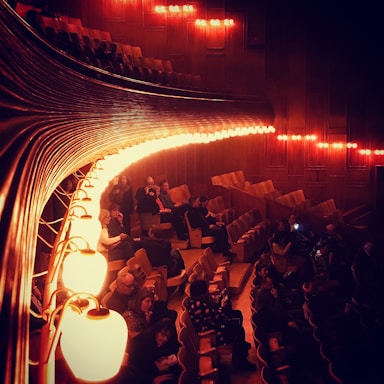 An auditorium with wooden interior features dimly lit, warm lights casting an amber glow. The seating area has several people scattered throughout, some are seated while others are standing or conversing. The curved lights run parallel to the rows of seats creating an elegant and intimate atmosphere.