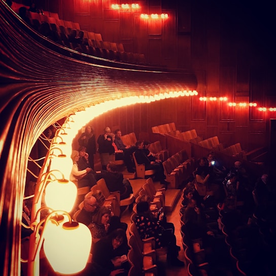 An auditorium with wooden interior features dimly lit, warm lights casting an amber glow. The seating area has several people scattered throughout, some are seated while others are standing or conversing. The curved lights run parallel to the rows of seats creating an elegant and intimate atmosphere.