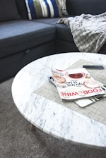 A marble round coffee table with a gray tablemat on top, holding two stacked magazines, a black remote control and a gray blanket draped over a dark gray sectional sofa with mixed throw pillows.