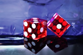 Two transparent red dice with white dots are positioned against a reflective surface and a blue-toned background. The lighting creates a dramatic contrast between the red and blue hues, enhancing the glossy appearance of the dice.