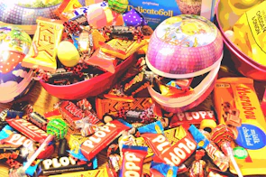 An assortment of colorful candy bars and sweets, including Twix, Plopp, and other chocolate treats, are scattered around and inside decorative Easter-themed containers. The packaging features bright and vibrant colors that create a playful and festive atmosphere.