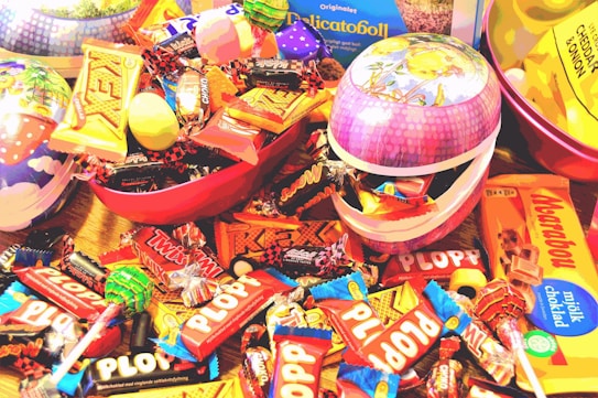 An assortment of colorful candy bars and sweets, including Twix, Plopp, and other chocolate treats, are scattered around and inside decorative Easter-themed containers. The packaging features bright and vibrant colors that create a playful and festive atmosphere.